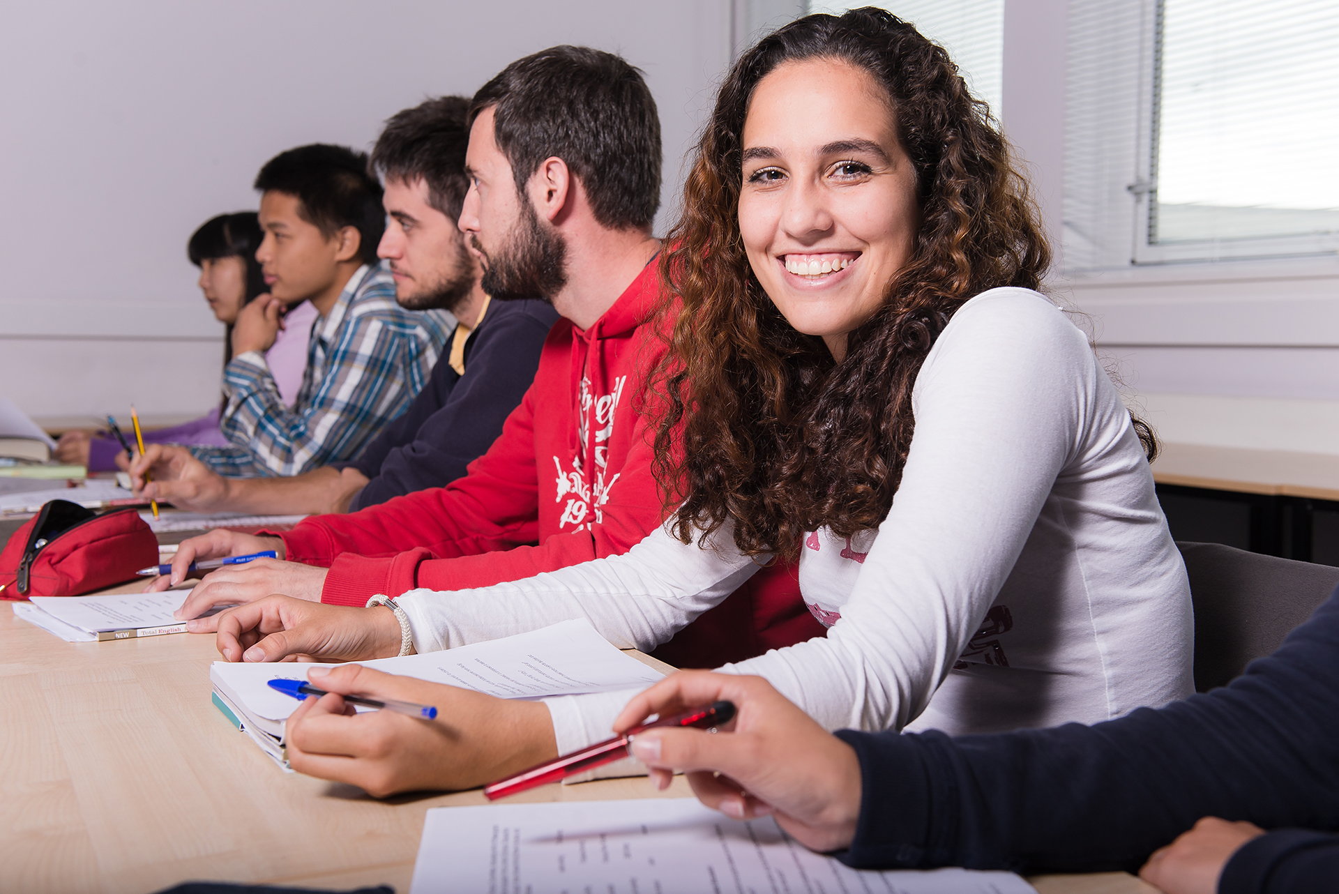 Smiling student is sitting at a desk with their peers, all holding pens and with notebooks in front of them.