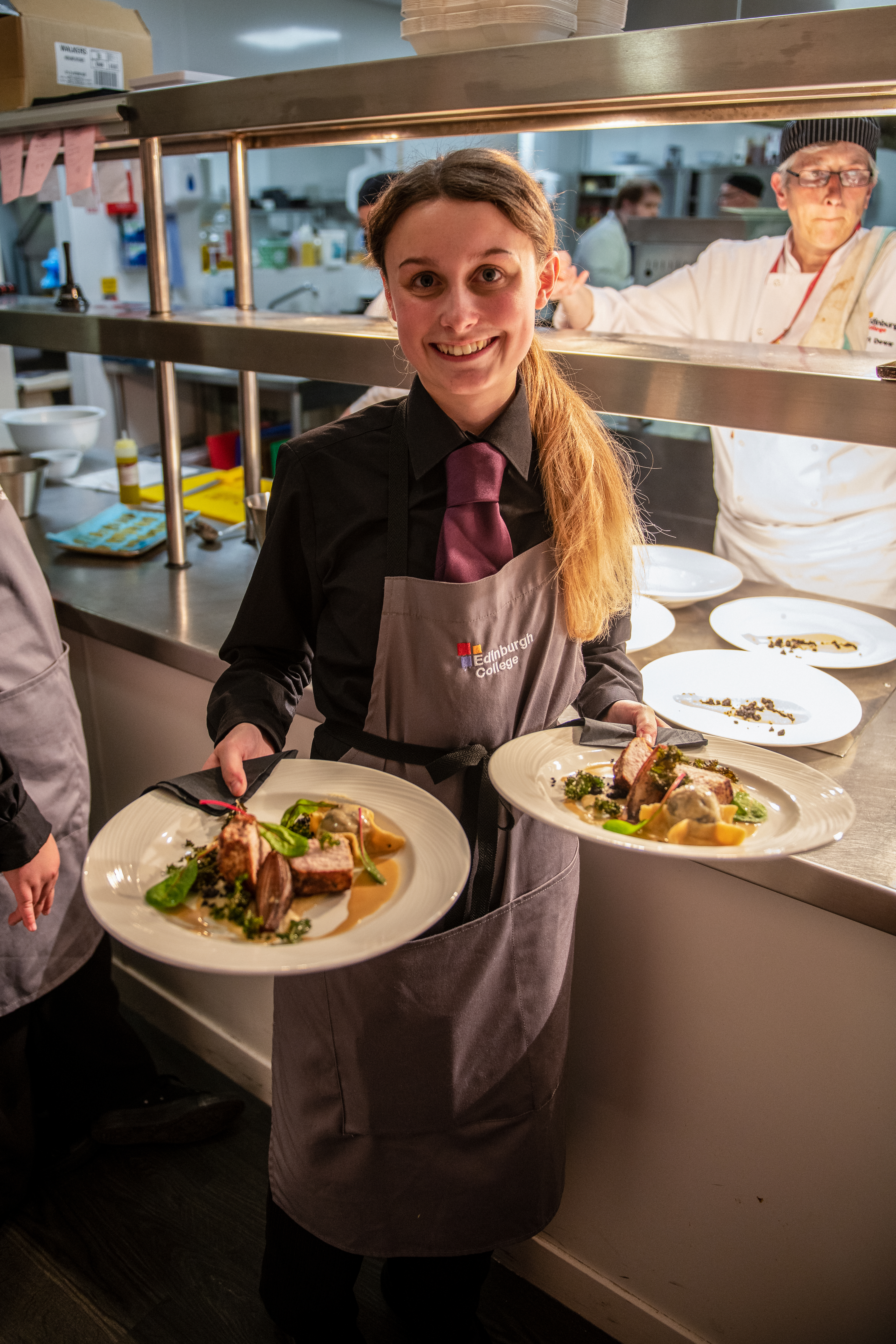 A waitress in a dark shirt and apron holds two elegantly plated dishes in a busy restaurant kitchen, with chefs working in the background.