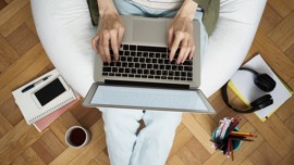 Shot from above, a person is sitting on a bean bag and typing on a laptop. The bean bag is surrounded by stationery, notebooks and a cup of cofee. 