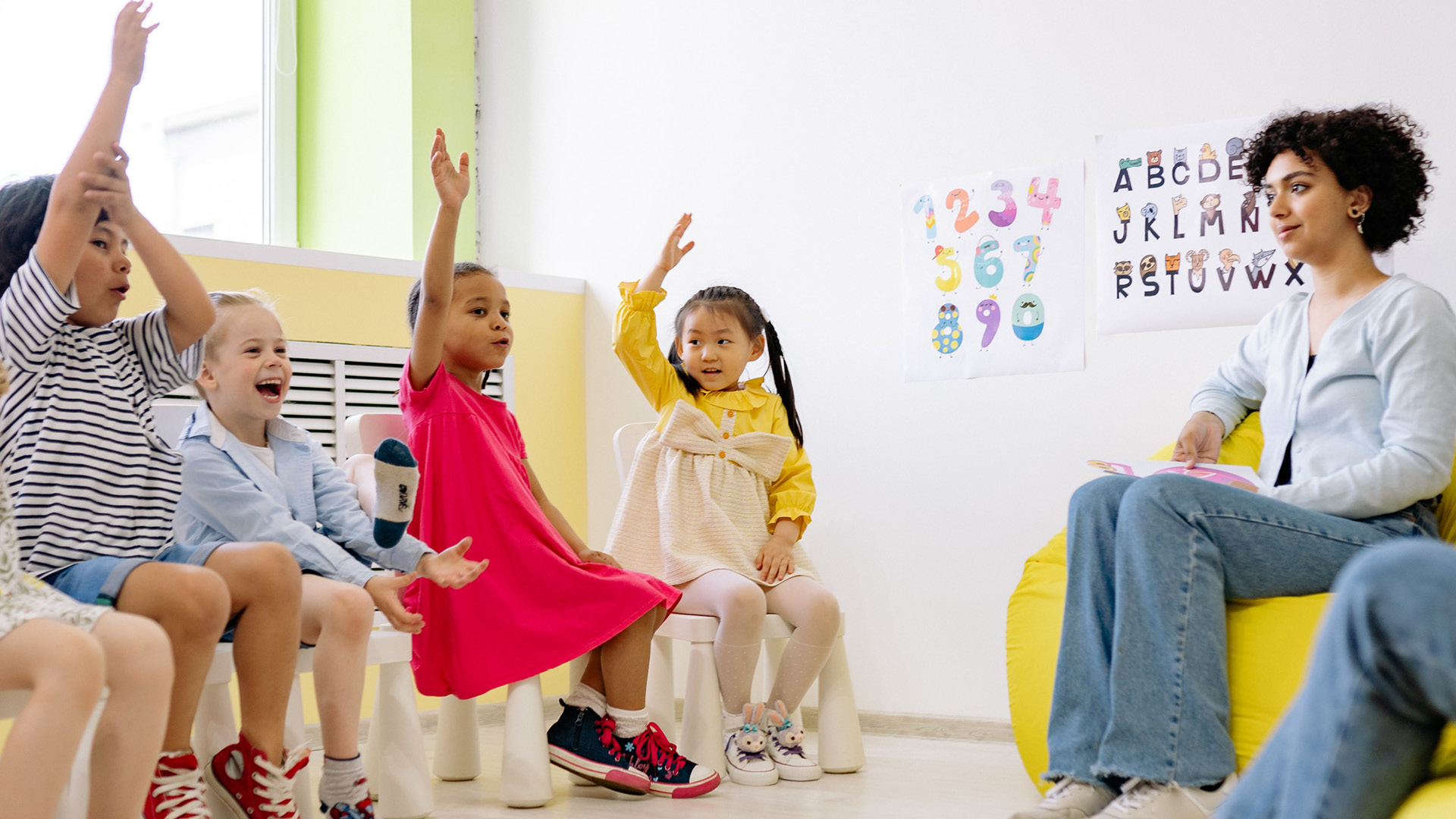 Group of young children holding up their hands in a nursery, with their teacher sitting near them.