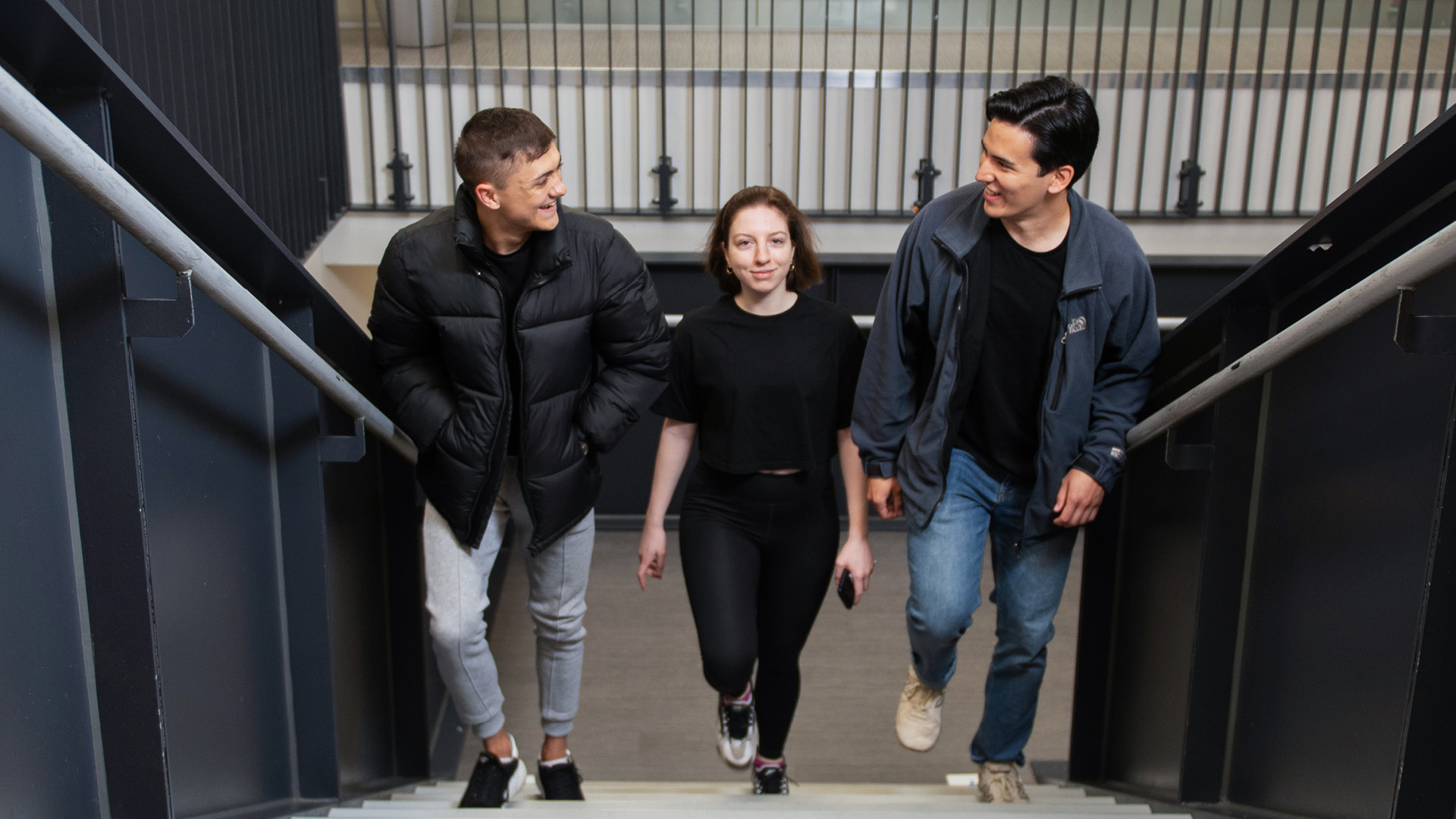 Three students talking and laughing with each other while walking up stairs at a college building.