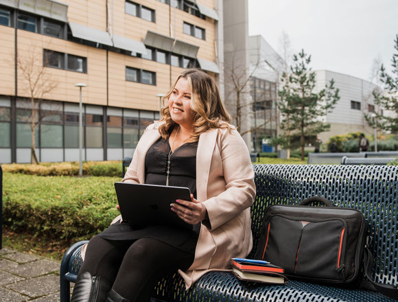 Student sitting at a bench outside a campus building, working on a tablet with their bag and books next to them.