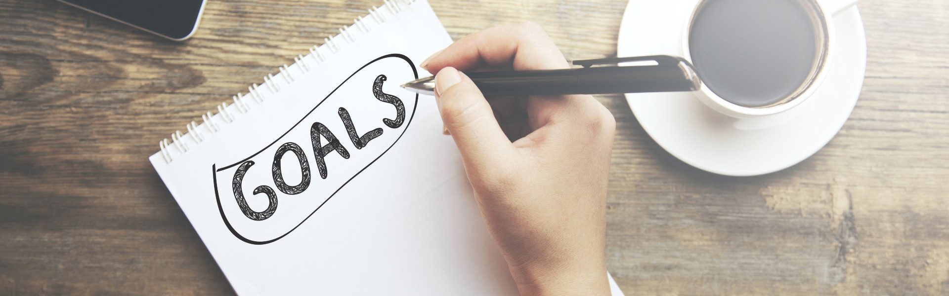 Notebook on a table surrounded by a mobile phone and cup of coffee. The word "Goals" is written on the notebook page.