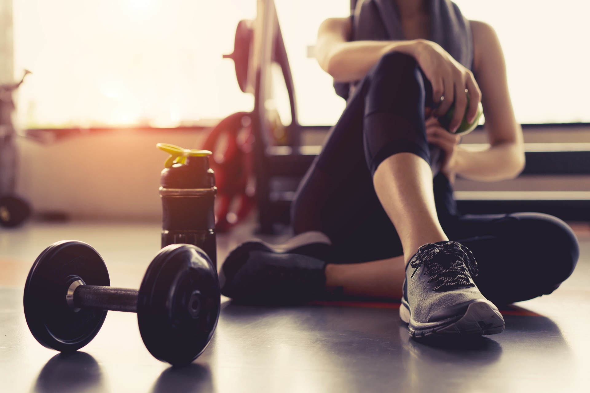 Person in fitness clothing sitting down in the gym with a water bottle and weights to their side.