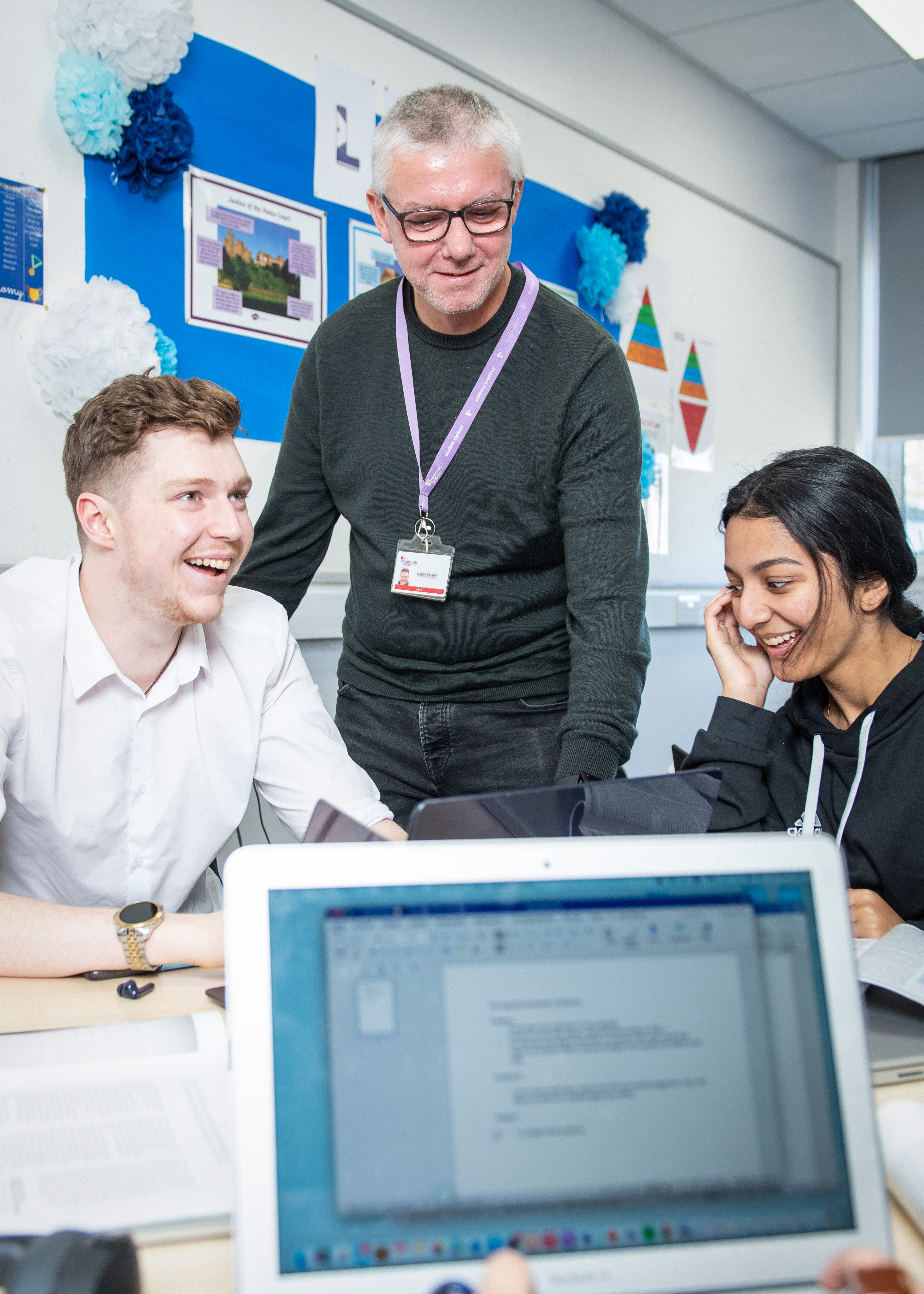 A teacher interacts with students in a classroom, while a laptop displays a text document on a desk. Classroom materials are visible.