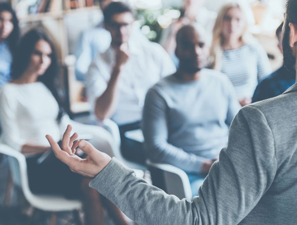 Person in a suit presenting in front of a group of people who are sitting down.