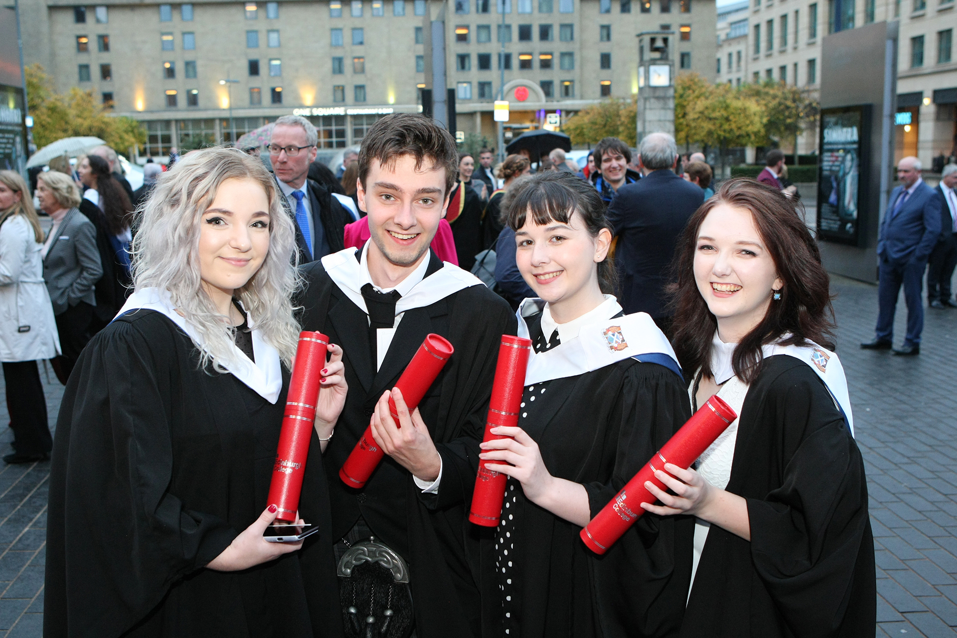 Four students at graduation, wearing black graduation gowns and holding red tubes with their scroll in.