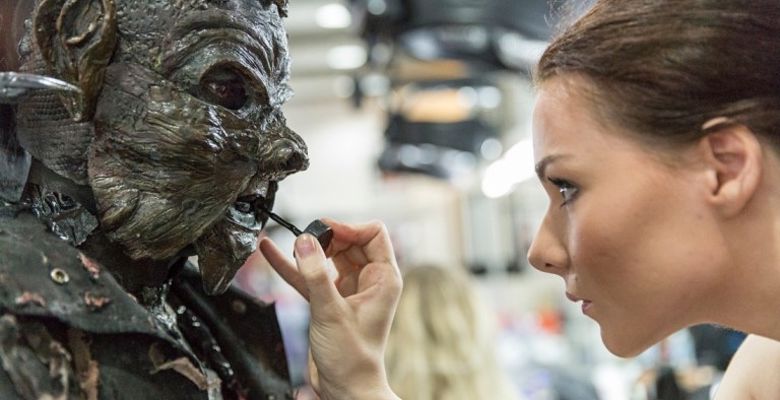 A character in an elaborate mask and textured makeup, resembling a creature sits while someone applies finishing touches with a brush.