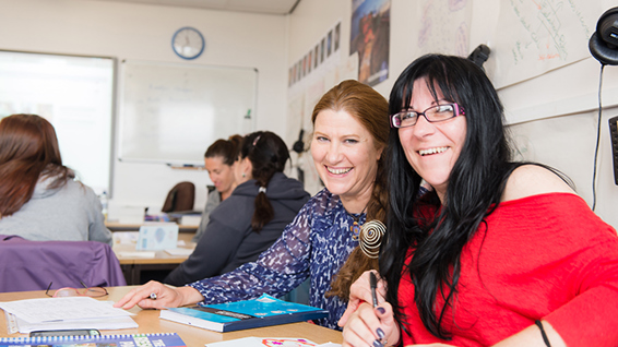 Two people pose together in a classroom setting filled with students, laptops, and classroom materials in the background.